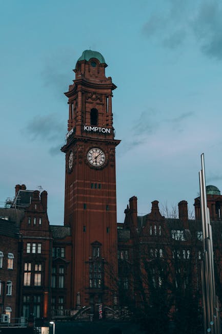 A tall red brick clock tower with a green dome at the top stands prominently against a cloudy sky, with the place name 'KIMPTON' illuminated on the tower. Surrounding the tower are historic buildings 